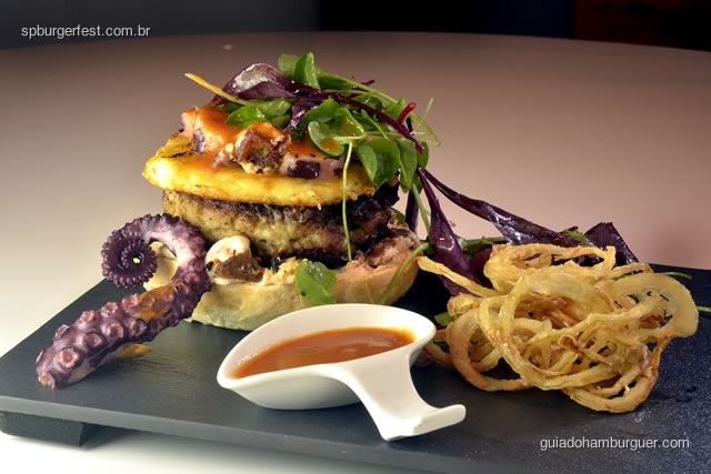 Hamburguer de pernil com polvo e gruyère - Base de pão caseiro de torresmo com maionese de páprica picante e limão siciliano, hambúrguer de pernil coberto com queijo gruyère e tentáculo de polvo. Servido com salada verde, onion ring e molho de tomate e polvo.  - SP Burger Fest 3ª edição